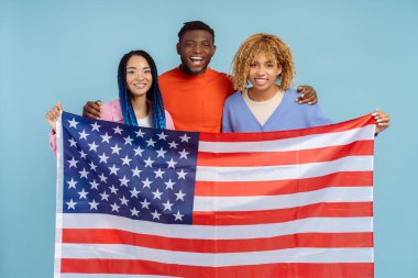 Amerikan bayrağı tutan mutlu Afro-Amerikalı arkadaşlar, mavi arka planda izole edilmiş kameraya bakıyorlar. Seçim sonuçları konsepti bekleniyor 