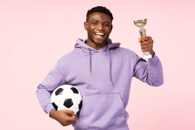 Zaferi kutlayan Afrikalı Amerikalı bir sporcunun portresi, elinde altın bir şampiyonluk kupası ve futbol topu, pembe arka planda izole edilmiş, kameraya bakıyor. 
