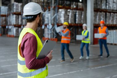 Başarılı bir orta yaşlı ustabaşı, elinde dijital tablet tutan sıkı bir şapka ve iş kıyafeti giyer, iş arkadaşlarının çalışmalarını izler, depoda çalışır. Kontrol kavramı, takım çalışması