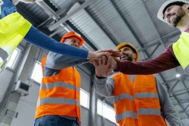 Bir grup fabrika işçisi, kasketli ve yelekli yöneticiler ellerini endüstri deposunda bir araya getiriyorlar. Takım çalışması konsepti