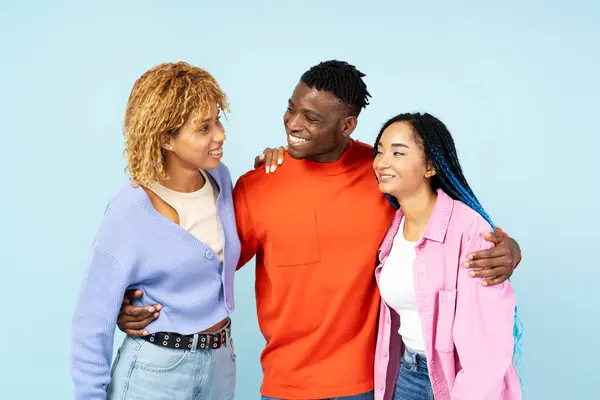 Young smiling friends wearing stylish clothing hugging, standing together isolated on blue background. Positive lifestyle, friendship concept