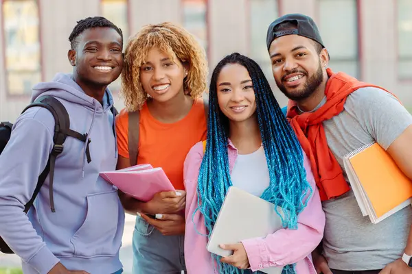 Group of smiling diverse African American friends in colorful casual clothes looking at camera near university campus on street. Happy students outdoors. Concept of education, communication, meeting
