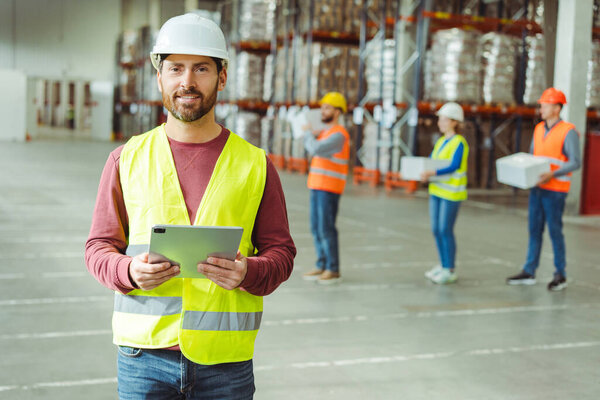 Smiling, attractive, bearded man, foreman wearing hard hat, holding digital tablet, supervises work of colleagues in warehouse. Concept of control, successful manager