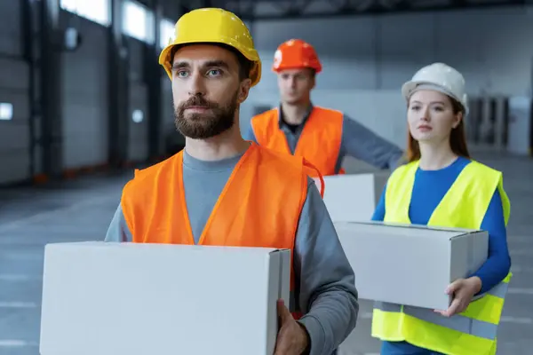 Group of workers, loaders, wearing hard hats and work uniforms, hold boxes, mockup looking away. Attractive bearded man carries box to warehouse. Concept of logistics, delivery