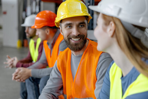 Positive colleagues wearing protective helmets and work wear, work in warehouse, talking during break. Smiling bearded worker, manager. Concept of teamwork, communication