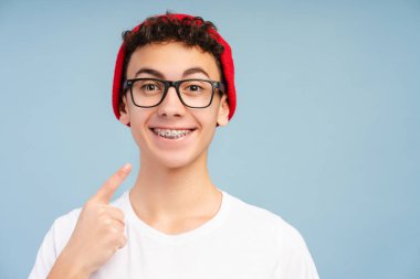Diş teli takan, şık kırmızı şapkalı ve gözlüklü gülümseyen, dişlerini gösteren, kameraya bakan, mavi arka planda tek başına duran genç bir çocuk. Diş sağlığı, tedavi, sağlık hizmetleri