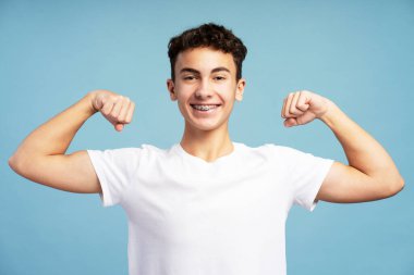 Gülümseyen, beyaz tişört giyen, kaslarını gösteren, mavi arka planda izole edilmiş kameraya bakan genç bir çocuk. Reklam kavramı