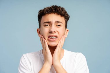 Üzgün genç çocuk yanağa dokunuyor, diş ağrısı mavi arka planda izole edilmiş. Diş teli takan mutsuz bir genç hasta. Diş sağlığı konsepti