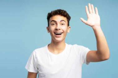 Yakışıklı, gülümseyen, heyecanlı el sallayan ve kameraya bakan çocuk. Yakışıklı genç, mavi arka planda izole edilmiş. Hoş geldin konsepti