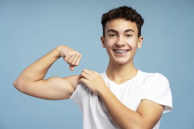 Gülümseyen, güçlü, şık beyaz tişört giyen, kaslarını gösteren, mavi arka planda izole edilmiş kameraya bakan genç bir çocuk. Reklam, spor konsepti