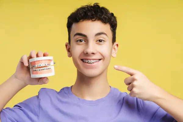 ハンサム 笑顔の少年 十代の若者は顎を保持し 黄色の背景に立っている間 カメラを見ている彼の歯の後ろに指を指しています 歯科医療 医療に関するコンセプト — ストック写真