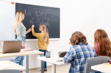 Öğretmen ona yardım ederken küçük bir kız tahtaya yazı yazıyor, sınıf arkadaşları sınıfın masalarında oturuyor. Eğitim konsepti. Okula geri dön.