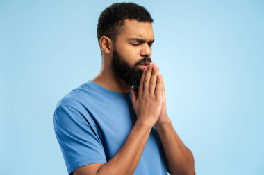 Sakin sakallı Afrikalı Amerikalı adam dua ederken kendine güveniyor, stüdyoda poz veriyor, mavi arka planda soyutlanmış. Halk yaşam tarzı konsepti