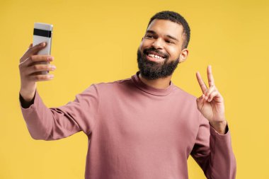 Akıllı telefondan fotoğraf çeken, sarı arka planda izole edilmiş zafer işareti gösteren pozitif sakallı Afro-Amerikan bir adam. Halk yaşam tarzı konsepti