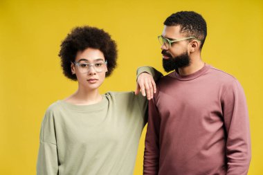 Gözlüklü, erkek omzuna yaslanan, stüdyoda poz veren, sarı arka planda izole edilmiş genç Afrikalı Amerikalı kadın. Yaşam tarzı insan konsepti