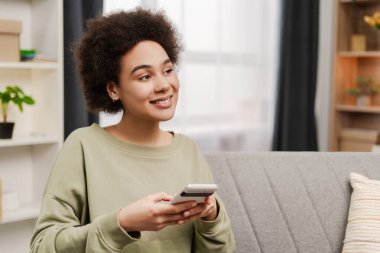 Evdeki kanepede otururken akıllı telefon kullanarak gülümseyen güzel Afro-Amerikan kadın, kopyalama alanı. Yaşam tarzı, teknoloji, insan kavramı
