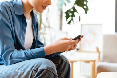 Neşeli, gülümseyen, cep telefonu tutan, akıllı telefon kullanan, video izleyen, evdeki rahat koltukta oturan, fotokopi çeken, odaklanmayı seçen genç bir kadın. Çevrimiçi teknoloji kavramı