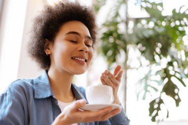 Gülümseyen, kıvırcık saçlı, elinde kahve fincanı olan ve fotokopi alanının güzel kokulu kokusunu alan Afro-Amerikan kadın. Kahvaltı konsepti, reklam.