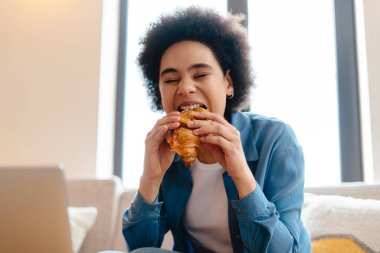 Kafede dizüstü bilgisayar kullanarak otururken gözleri kapalı taze kruvasan yiyen mutlu, genç Afrikalı Amerikalı hippi kadın. Yemek molası, öğle yemeği.