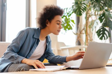 Gülümseyen Afro-Amerikan kadın, serbest çalışan, kanepede oturan, dizüstü bilgisayar kullanan, uzaktan kumandalı, online eğitim alan. Sosyal medya kavramı