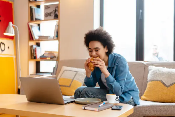 Portrait of young African American woman, freelancer, stylish hipster using laptop, eating delicious croissant while sitting on comfortable sofa in modern cafe. Concept of remote job, lunch, break