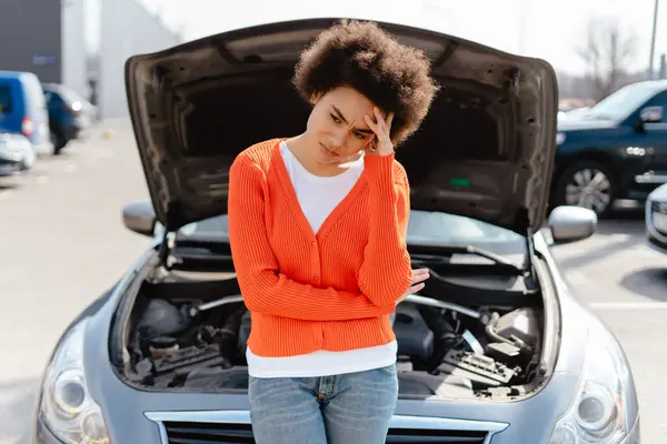 Upset, sad African American woman with curly hair standing by open hood of broken car waiting for help on the street. Concept of car insurance, car repair