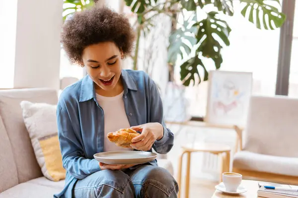 カフェで快適なソファーに座っている間 空腹で興奮し 魅力的なアフリカ系アメリカ人女性がおいしいクロワッサンを食べます 食べ物 朝のルーチン スナックのコンセプト ストック画像