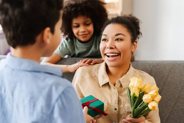 驚いたアフリカ系アメリカ人女性は子供たちから贈り物を受け取り 自宅でたくさんの花とギフトボックスを持っていました 母の日のコンセプト お祝い ストックフォト