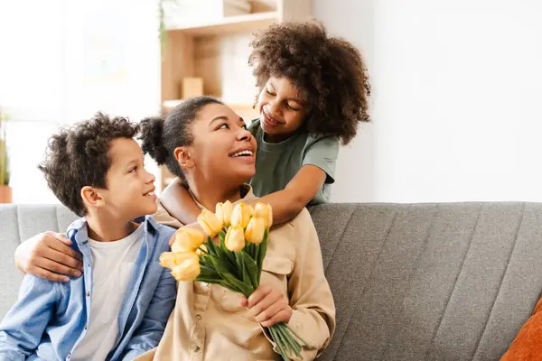 幸せな若いアフリカ系アメリカ人女性は 子供たちと祝いながら 自宅でソファーに座りながら たくさんの花を持っています 母の日のコンセプト お祝い ストックフォト