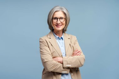 Smiling senior business woman wearing eyeglasses looking at camera, posing arms crossed isolated on blue background. Portrait of confident gray haired politician. Successful business, career clipart
