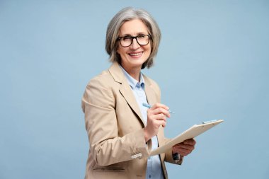 Yakışıklı, çerçeveli gözlük takan, not defterine notlar alan, proje planlarken kameraya bakan, yapılacaklar listesi yazan, programları analiz eden, mavi arka planda izole edilmiş son sınıf öğrencisi bir kadın.