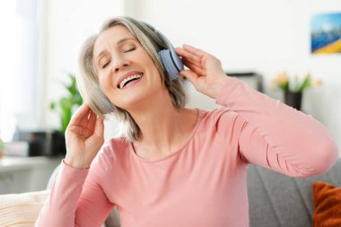 Çekici, mutlu, son sınıf öğrencisi, kablosuz kulaklıkla müzik dinleyen, evdeki kanepede rahat bir şekilde oturan bir kadın. Teknoloji, pozitif yaşam tarzı konsepti 