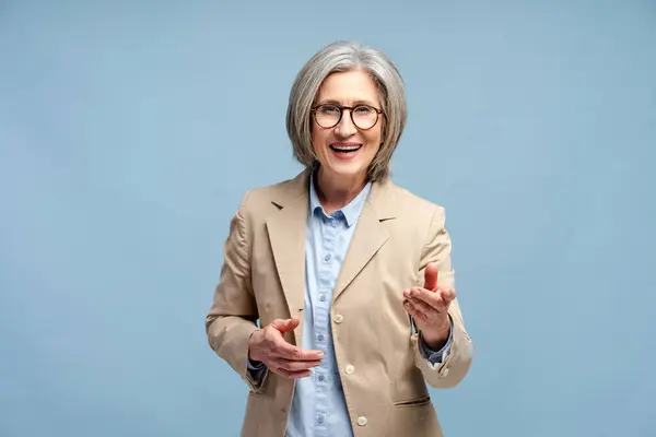 青い背景に隔離された指を指す眼鏡とスタイリッシュなスーツを着た60歳の女性の笑顔 楽しいコンセプトを持つこと ストック写真