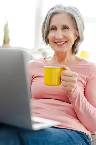 ラップトップコンピュータを使用して幸せなシニアの女性の垂直な眺め 自宅で快適なソファーに座ってコーヒーカップを保持します 自宅で働くグレイハイレッドビジネスマン ストックフォト