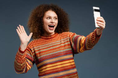 Kıvırcık saçlı mutlu bir kadın, günlük kıyafetler giyen, cep telefonu tutan, video kaydeden, selfie çeken, el sallayan, gri arka planda tek başına duran bir blogcu. Çevrimiçi teknoloji kavramı