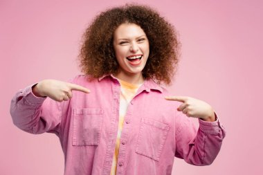 Kıvırcık saçlı, neşeli, sevimli bir kadın pembe arka planda izole bir şekilde kameraya bakarken kendini işaret ediyor. Alışveriş konsepti, reklam