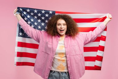 Kıvırcık saçlı, elinde Amerikan bayrağı olan neşeli bir kadın seçim sonuçlarını bekliyor, kameraya bakıyor, pembe arka planda tek başına duruyor. Başarılı ABD başkanlık seçimi kavramı 2024