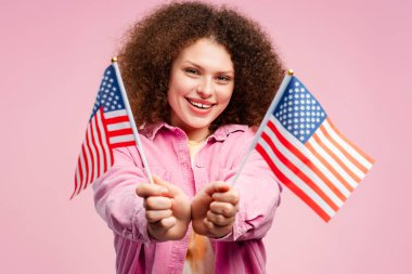 Heyecanlı, kıvırcık saçlı, destekçisi olan, Amerikan bayrağını tutan, pembe arka planda izole bir şekilde duran genç bir kadın. 2024 ABD başkanlık seçimi kavramı