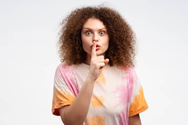 Çekici, kıvırcık saçlı, sessiz bir jest yapan, parmaklarını ağzının kenarında tutan, kameraya bakan, beyaz arka planda yalnız duran genç bir kadın. Gizli konsept