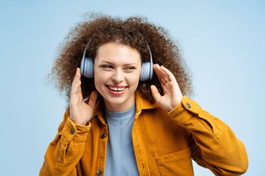 Kıvırcık saçlı, şık sarı ceketli, kablosuz kulaklıkla müzik dinleyen, mavi arka planda tek başına duran çekici genç bir kadının portresi. Teknoloji konsepti