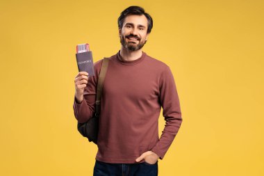 Sırt çantasında pasaport, bilet taşıyan mutlu bir gencin portresi, sarı arka planda izole edilmiş. Turizm ve tatil kavramı