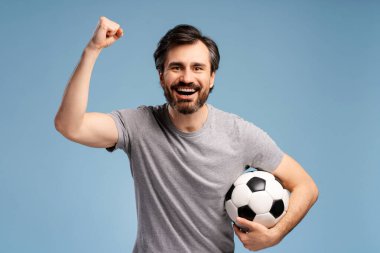 Yakışıklı genç futbolcu, izole edilmiş mavi arka planda, yüzünde mutlu bir ifade var, topu tutuyor. Spor, tezahürat, kutlama kavramı