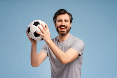 Genç, yakışıklı, pozitif adam futbol topunu mavi arka planda izole bir şekilde kameraya bakarak tutuyor. Spor, tezahürat, kutlama kavramı