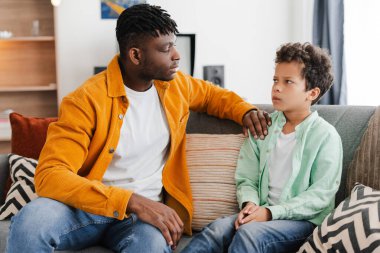 Genç Afrikalı Amerikalı bir adam rahat bir odada rahat bir kanepede otururken küçük bir oğluyla konuşuyor. İlişki konsepti, ebeveynlik, iletişim