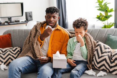 Üzgün, hasta, Afrikalı bir baba ve küçük bir oğul, burunları akmış, üşümüş, virüs kapmış, peçete tutuyorlar, evdeki rahat kanepede birlikte oturuyorlar. Ebeveynlik, sağlık, tedavi kavramı