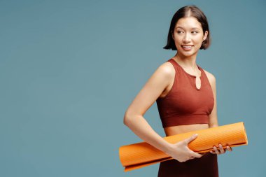 Mutlu gülümseyen kadın, kaslı güçlü estetik vücudu başka tarafa bakıyor, fitness yoga minderini tutuyor, mavi arka planda izole edilmiş.