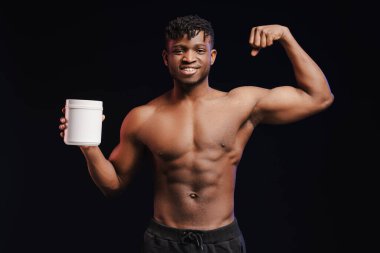 Gülümseyen Afrikalı bir adam, vücut geliştirici, çıplak gövdeli bir sporcu elinde beyaz şişe maketi olan, pazularını gösteren, kameraya bakan, siyah arka planda tek başına duran, spor besleyicisi, takviye.