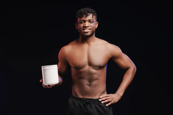 stock image Handsome, smiling, african american man, pumped athlete with bare torso, holding white bottle, mockup, copy space, standing isolated on black background. Concept of sports nutrition, supplement