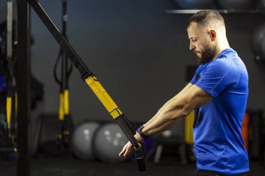Attractive, handsome man, strong trainer, athlete doing functional training, holding TRX equipment, standing in modern gym. Concept of sport, healthy lifestyle