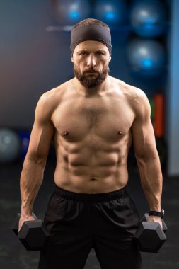 Brutal, sexy, serious man, athlete with naked pumped torso holding dumbbells, training in gym, looking at camera. Sports concept